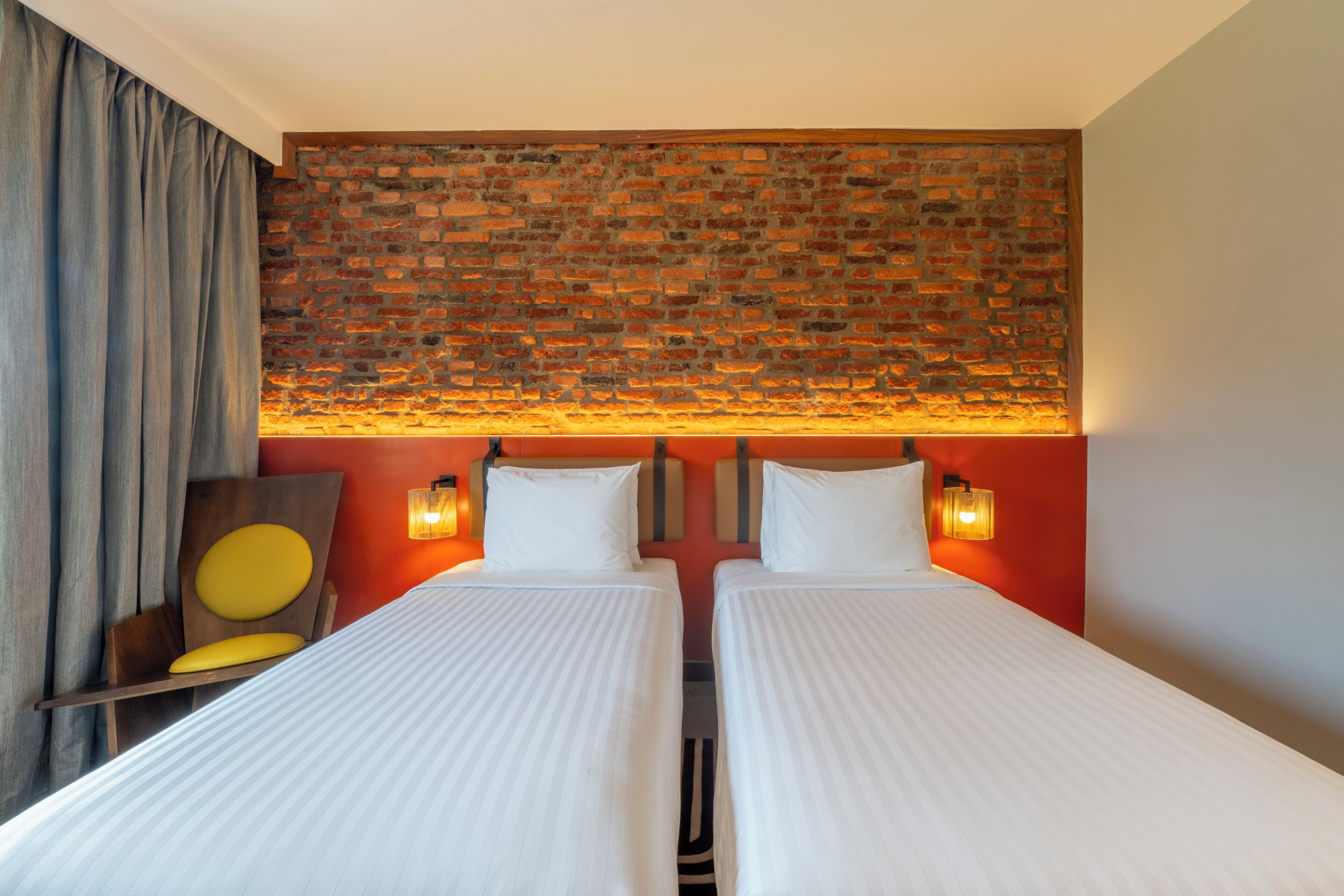 A hotel room with two beds, a brick wall headboard, a wooden chair with a yellow cushion, and a red accent wall.