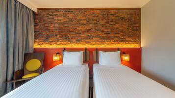A hotel room with two beds, a brick wall headboard, a wooden chair with a yellow cushion, and a red accent wall.
