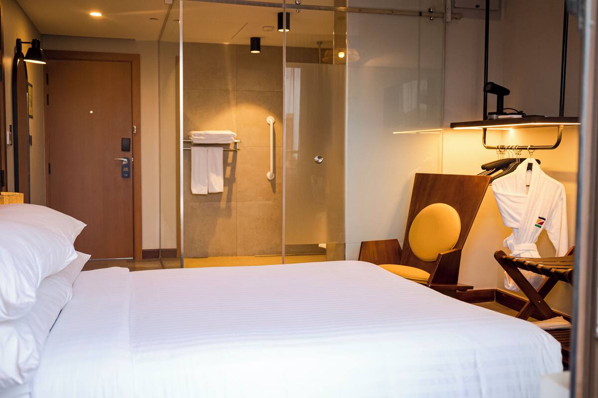 A hotel room with a large bed, a glass-enclosed shower, a wooden chair, and a robe hanging on a rack.