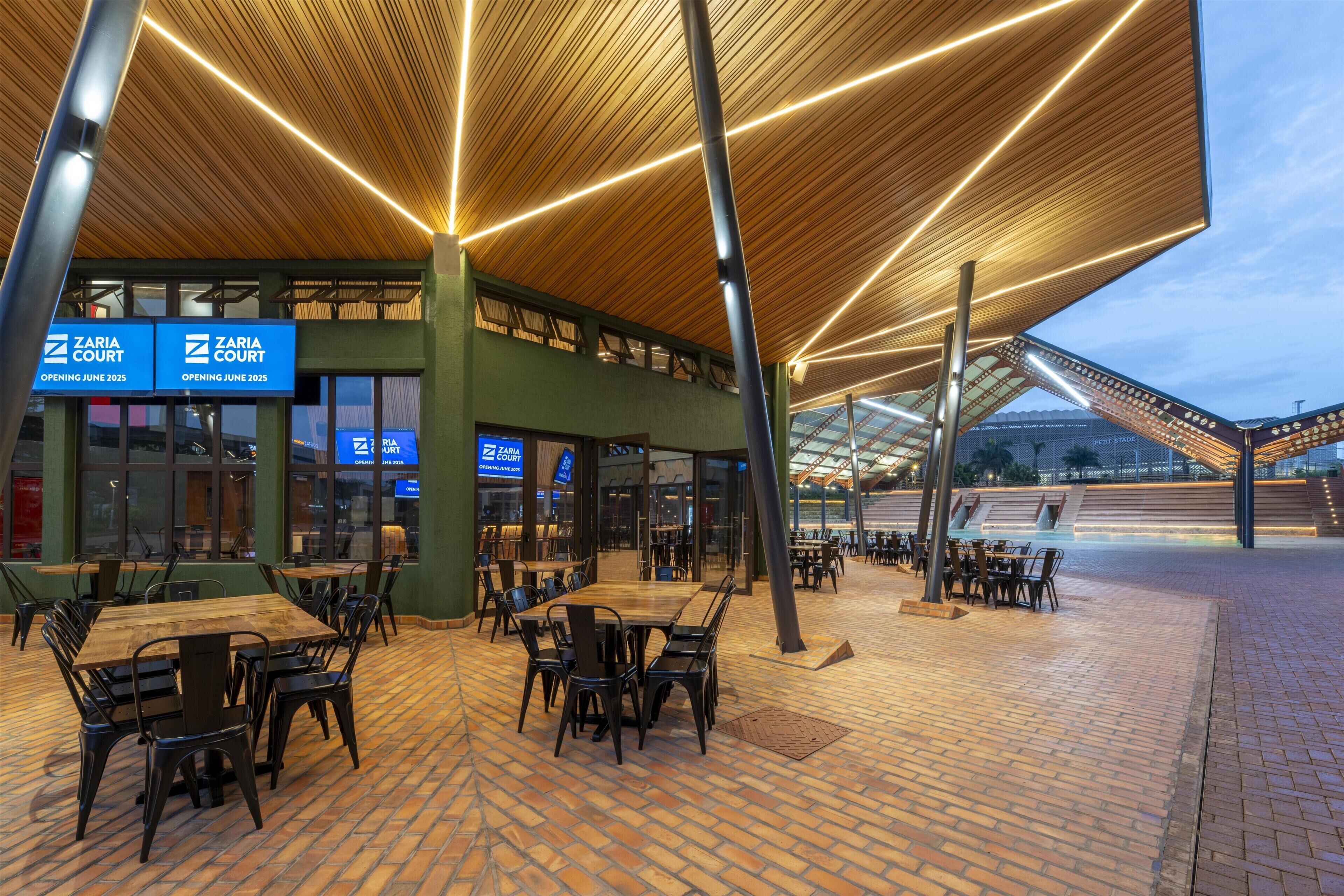 An outdoor seating area with tables and chairs, a covered section with a wooden ceiling, and a building with 'ZARIA COURT' signage.