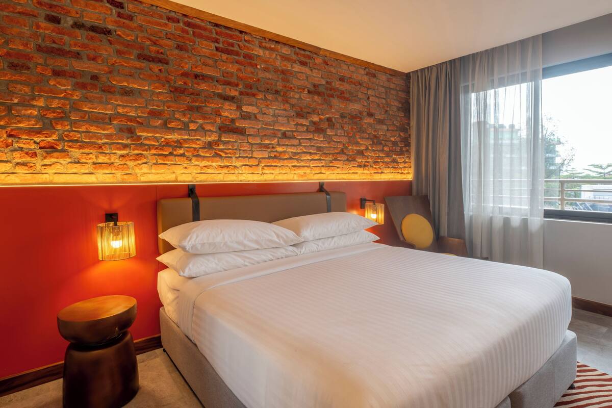 A hotel room with a brick wall, a large bed with white linens, a wooden stool, and a nightstand with a lamp.