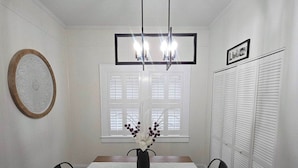 A dining room with a wooden table, four chairs, a chandelier, a window with shutters, and a wall-mounted picture frame.