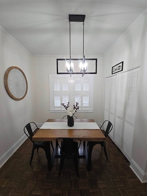 A dining room with a wooden table, four chairs, a chandelier, a window with shutters, and a wall-mounted picture frame.