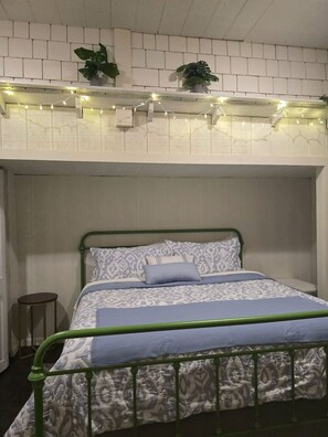 A bed with a patterned bedspread and pillows, a green metal frame, a small round table, and a wall with a brick pattern and string lights.