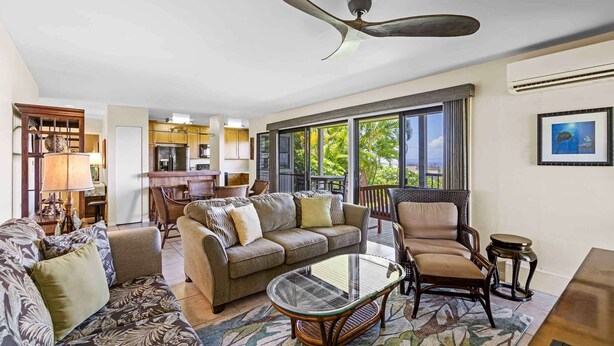 A spacious living room with a sofa, armchairs, and a coffee table. There is a ceiling fan, large windows, and a kitchen in the background.