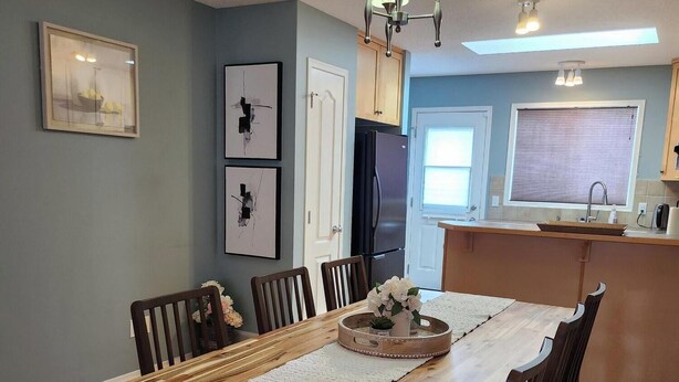 A dining area with a wooden table, chairs, and a chandelier. There is a kitchen in the background with a refrigerator and a sink.
