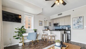 A modern apartment with a living room, kitchen, and dining area. It features a flat-screen TV, a ceiling fan, and a large window.