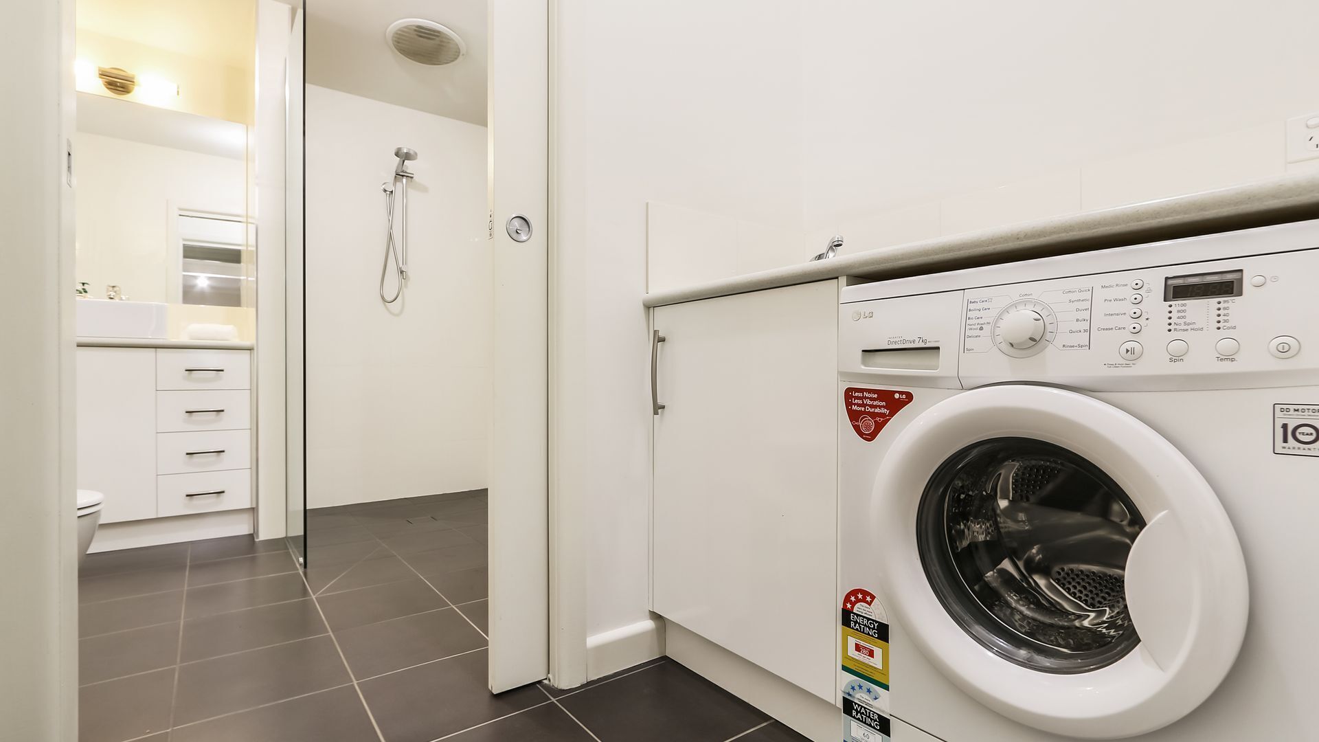 A laundry room with a front-loading washing machine, a shower, and a white bathroom with a sink and drawers.