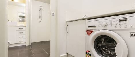 A laundry room with a front-loading washing machine, a shower, and a white bathroom with a sink and drawers.