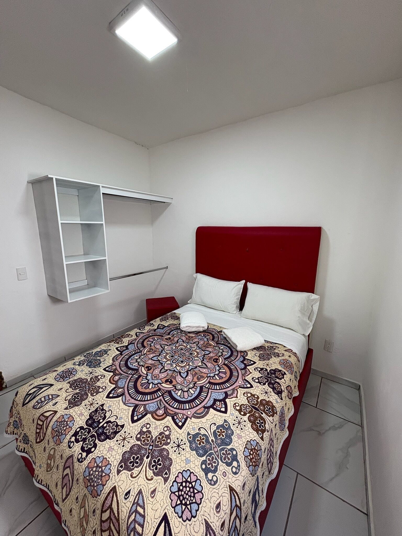 A bedroom with a bed, a red headboard, a patterned bedspread, a white bedside table, and a white shelf unit.