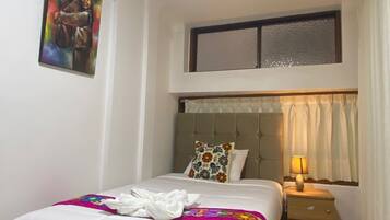 Un dormitorio ordenado con cama, una mesita de noche con lámpara, una ventana con cortinas y un cuadro en la pared.