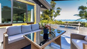 A modern outdoor seating area with wicker furniture, a glass-top table, and a view of the ocean. - Glass Horizon - Rutherglen Holiday Home (Ruthherglen)