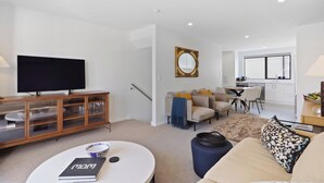 A modern living room with a flat-screen TV, a dining area with a table and chairs, a kitchen with a window, and a cozy seating area with a sofa and cushions. - Modern Central Apartment - Christchurch Apartment (Christchurch)