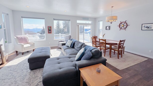 A spacious living room with a sectional sofa, a dining table with chairs, and large windows offering a view of the outdoors. - 3-Bedroom House in Tranquil Homer, Alaska. Perfect Family Retreat. (Homer)