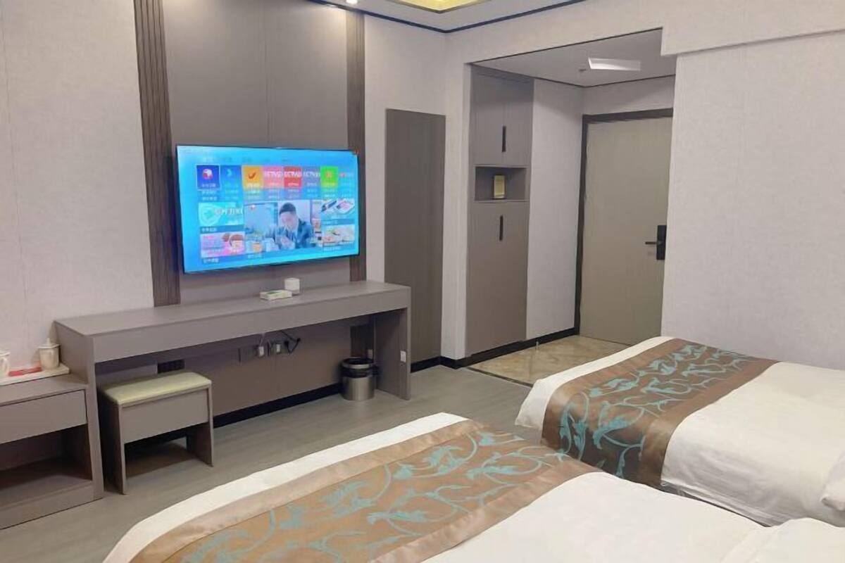 A hotel room with a TV on a stand, a desk, a bench, two beds with patterned bedspreads, and a wardrobe.