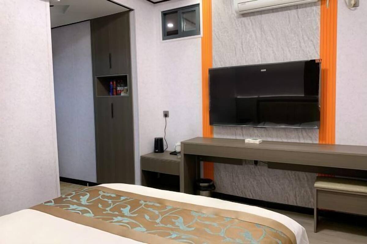 A hotel room with a bed, a desk, a television, a wardrobe, and a small shelf with items.