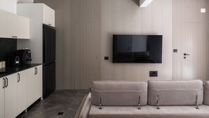 A modern kitchen with a flat-screen TV mounted on the wall, a sofa in the seating area, and a coffee machine on the counter.