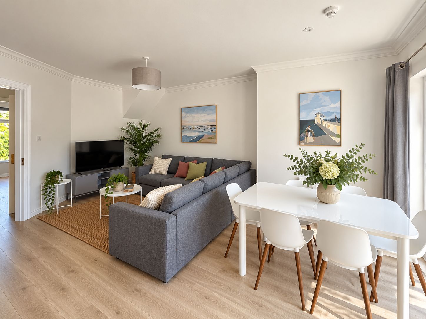 A modern living room with a grey sofa, a flat-screen TV, a dining table with chairs, and two paintings on the wall.