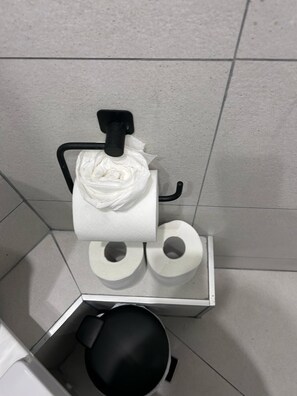 A corner of a bathroom with a toilet paper holder, two rolls of toilet paper, and a black trash bin.