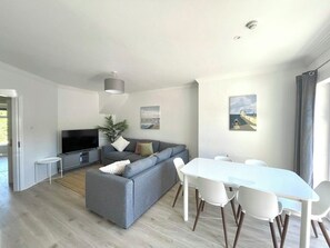 Un salón moderno con un sofá gris, un televisor de pantalla plana, una mesa de comedor con sillas blancas y un ventanal con cortinas.