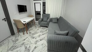 Ein kleiner, moderner Wohnraum mit einem grauen Sofa, einem kleinen Tisch und einem einzelnen Sessel. Ein Fernseher ist an der Wand befestigt.
