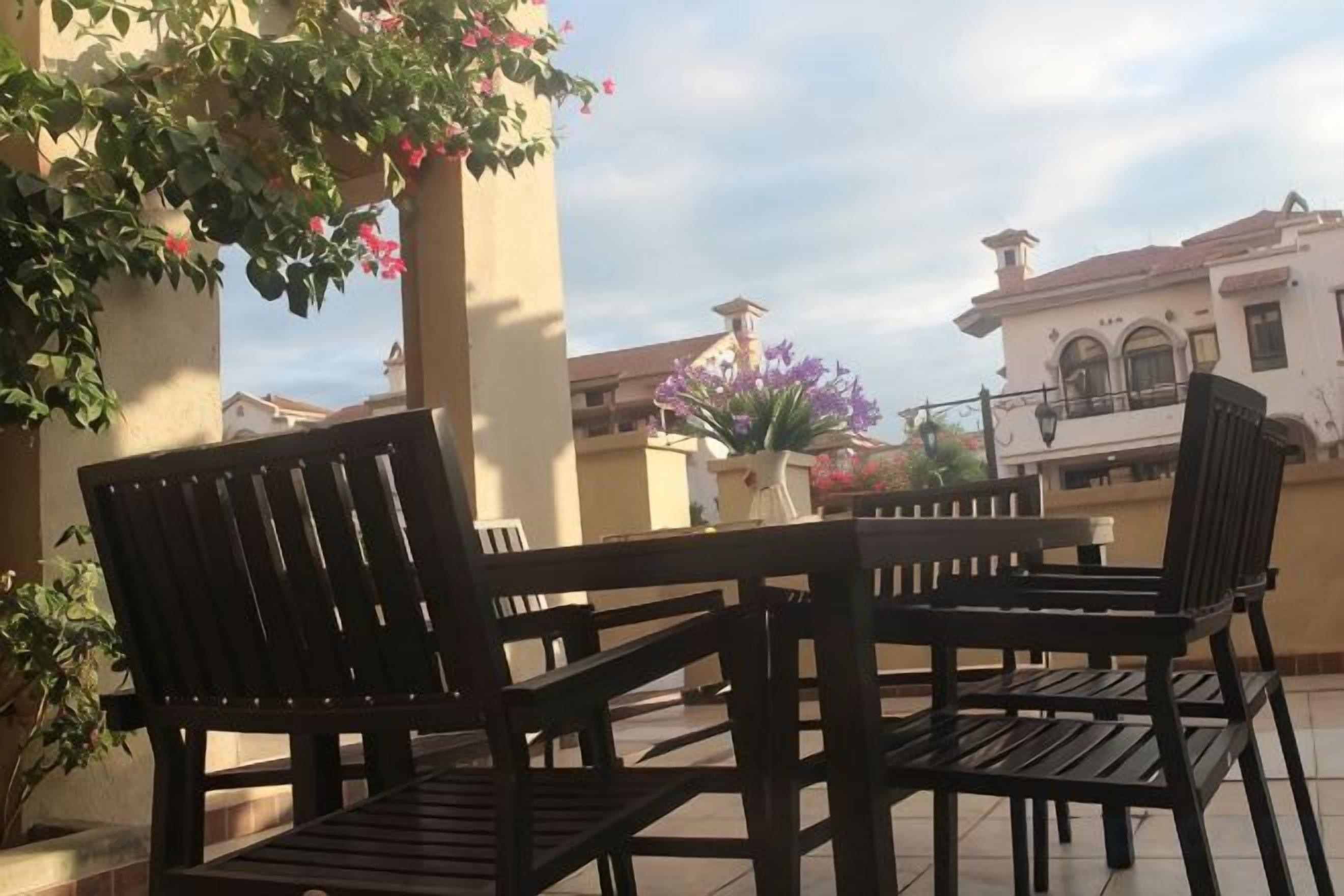 An outdoor patio with wooden chairs and a table, surrounded by potted plants and a building with a tiled roof.
