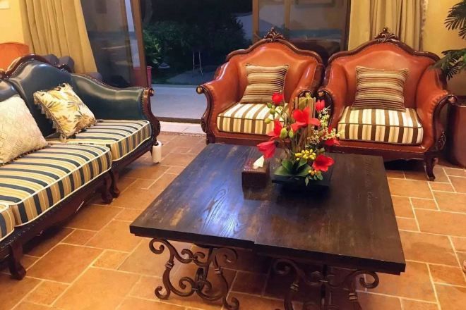 Een kamer met een donker houten bijzettafeltje, twee gestoffeerde banken met gestreepte kussens, en een vaas met bloemen.