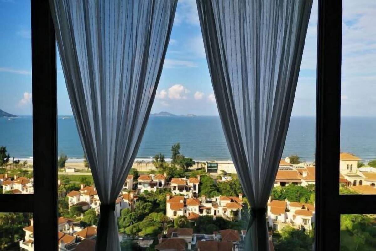 Blick durch Vorhänge auf eine Küstenstadt mit Häusern und dem Meer.