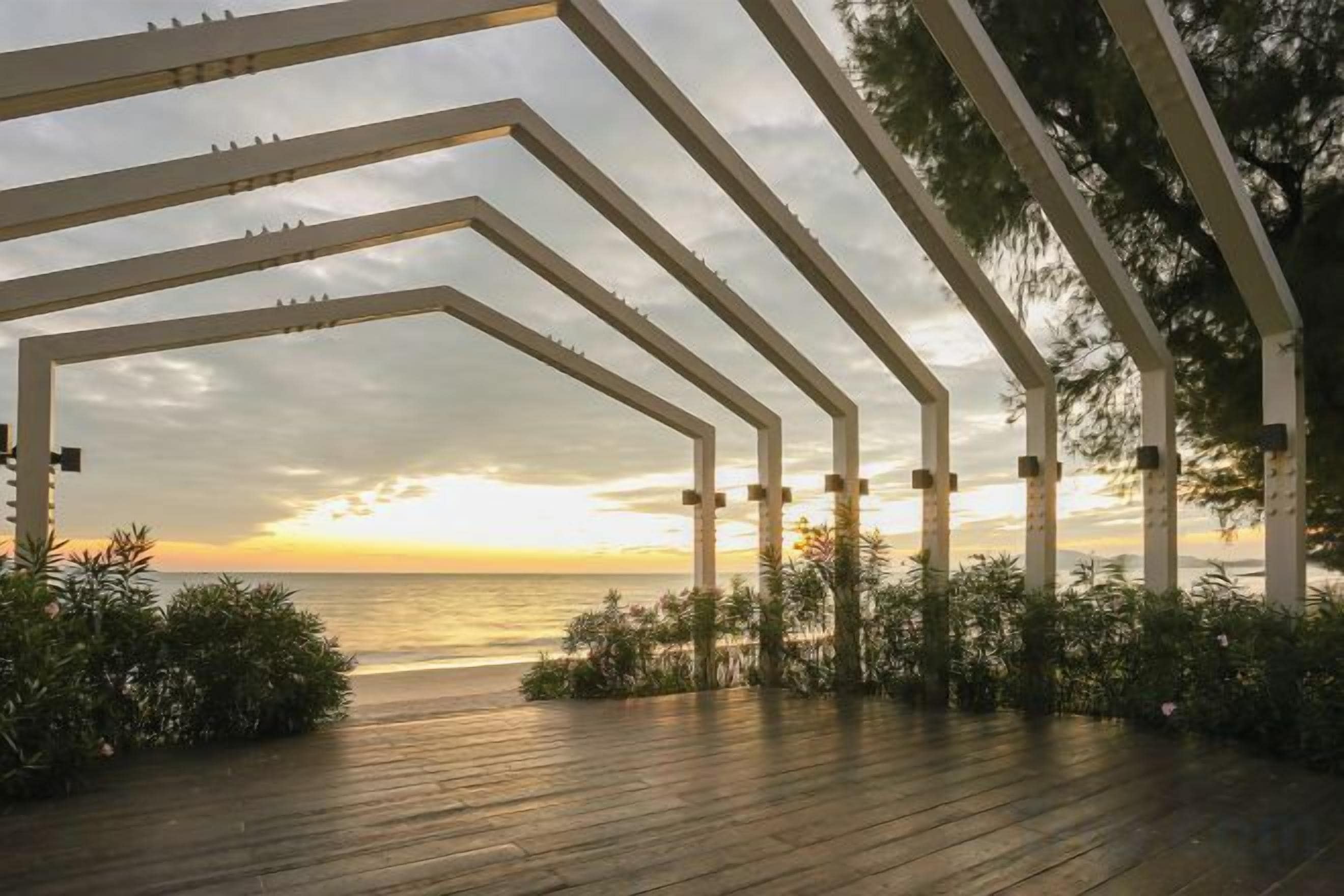 Una terraza de madera con estructura de pérgola, con vistas a una masa de agua durante el atardecer.