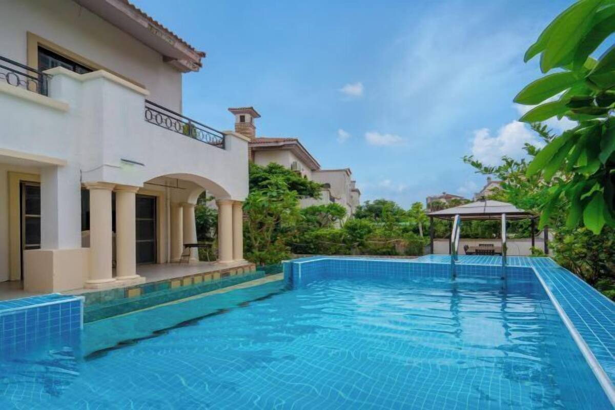 Ein Pool mit gefliestem Rand, eine überdachte Terrasse und ein Gebäude mit Säulen und einem Balkon.