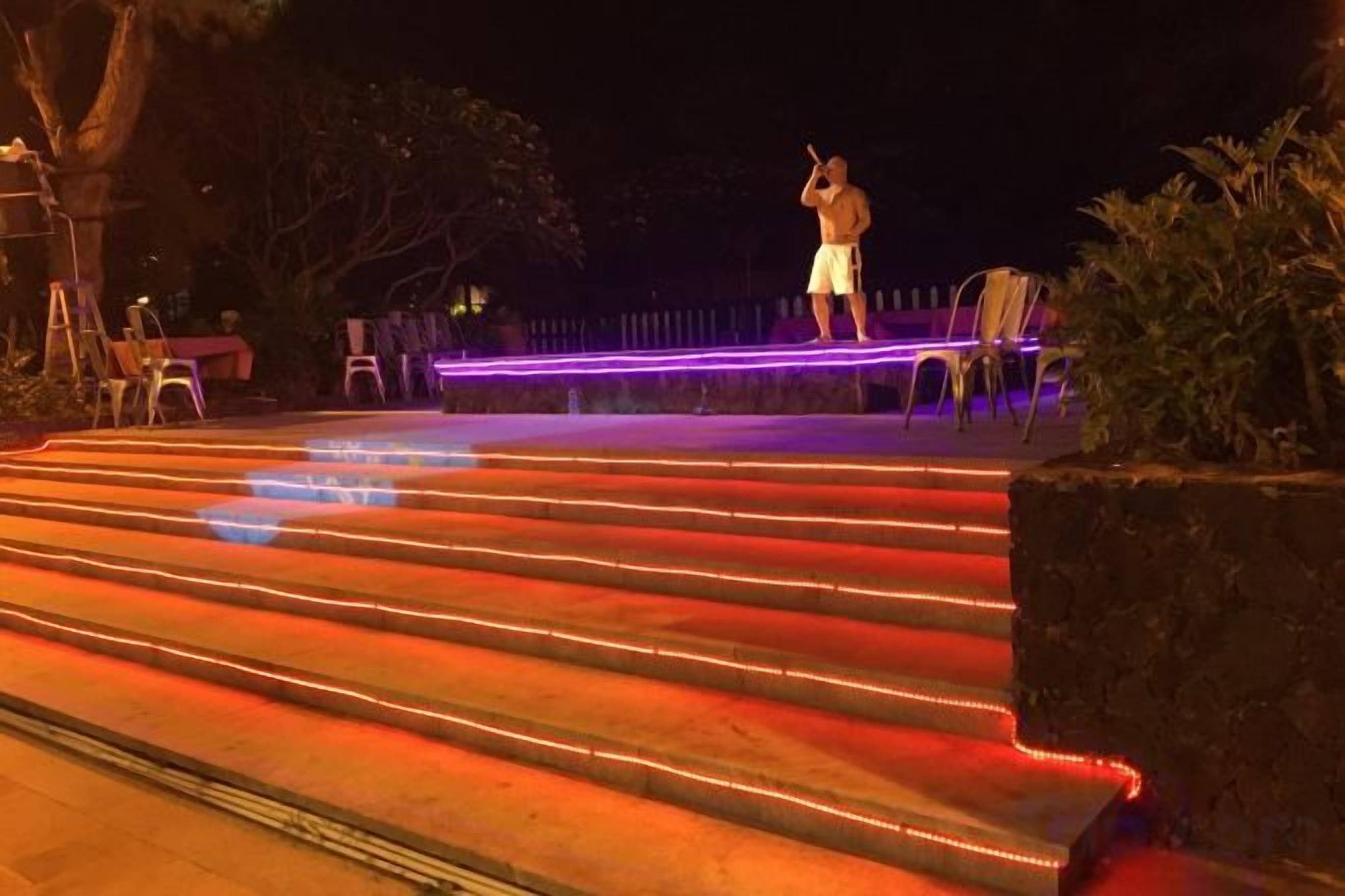 Una persona parada en unas escaleras exteriores con un escenario, rodeada de sillas y plantas.