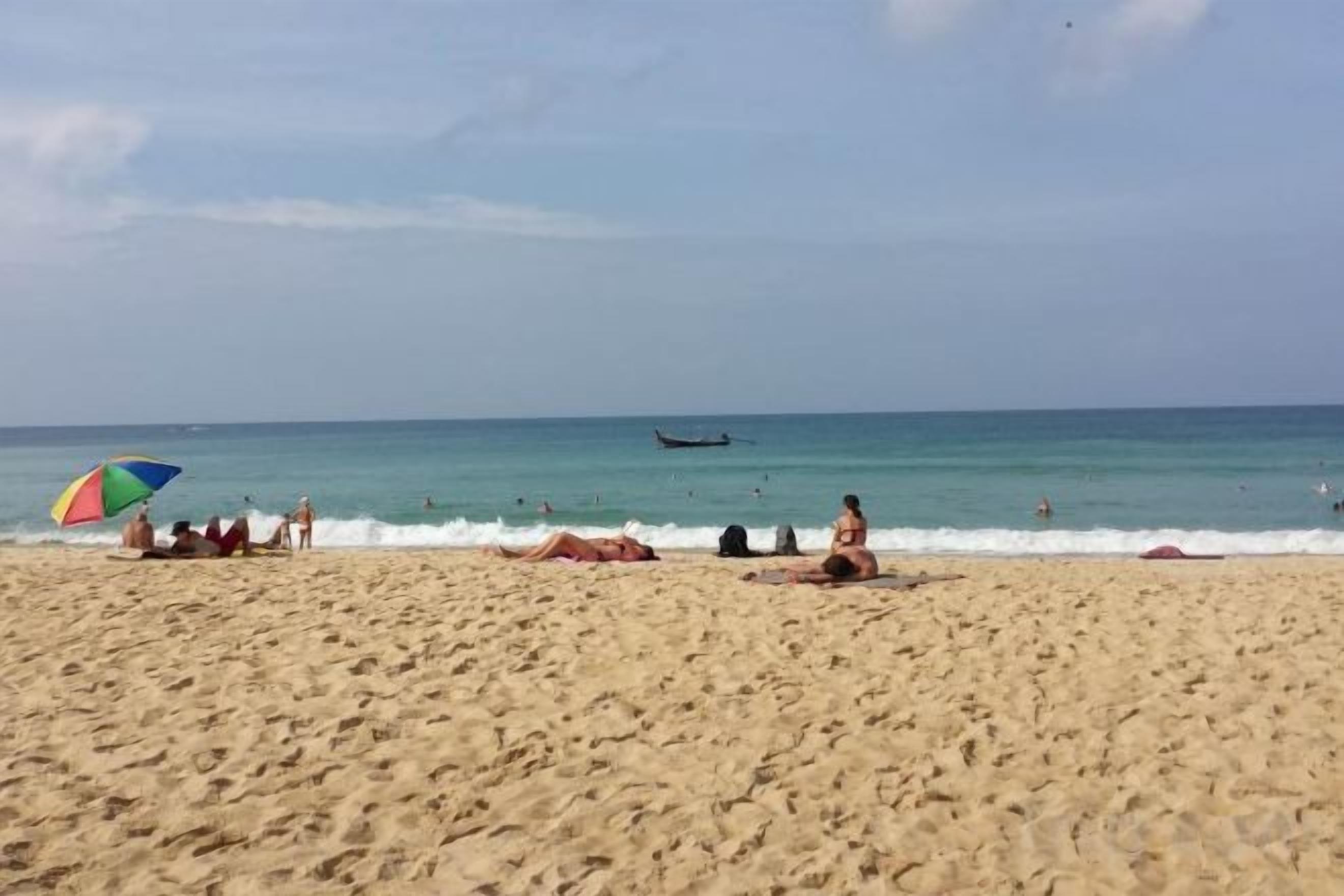 A beach with people sunbathing, a colorful umbrella, and a plane in the distance.