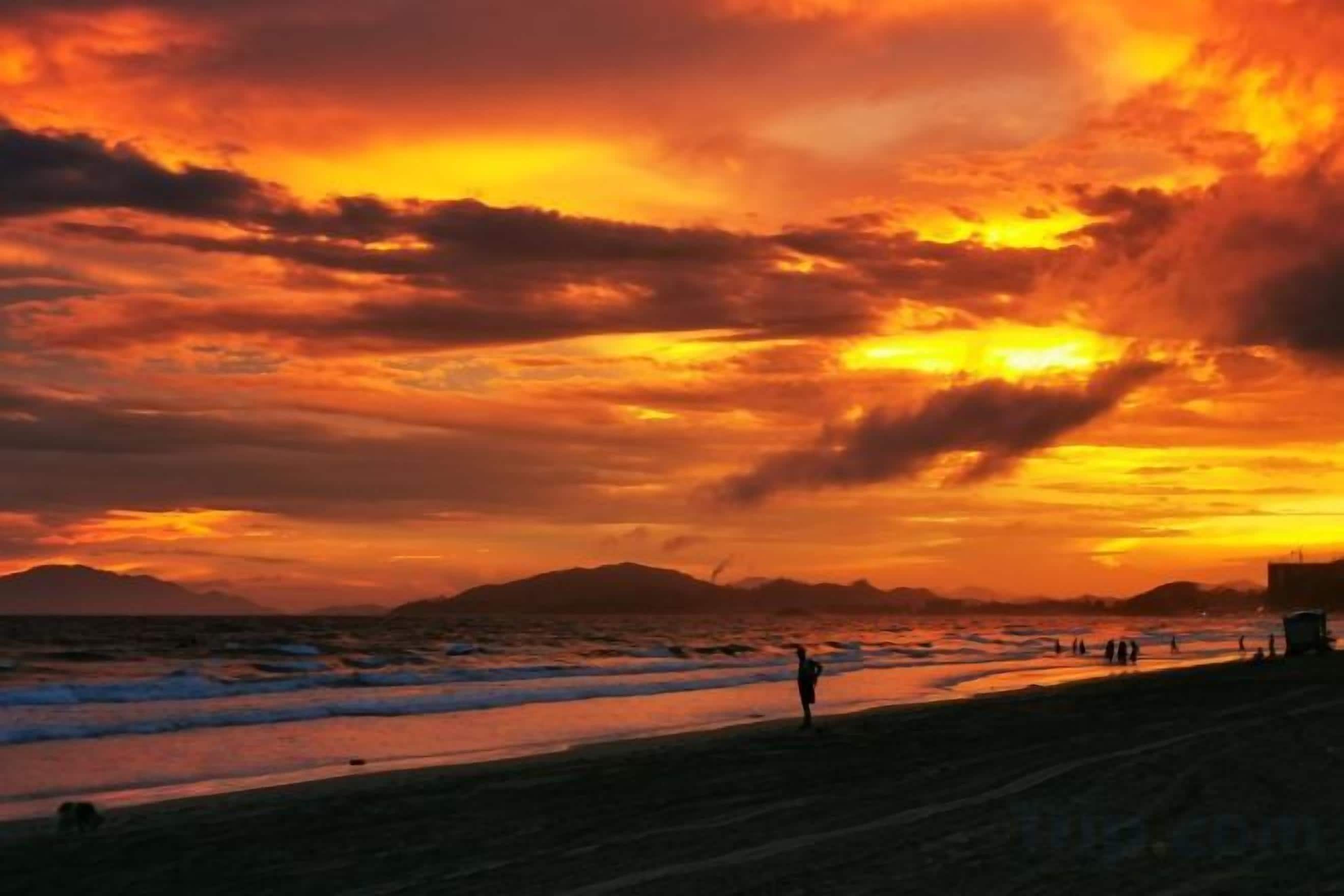 Une scène de plage au coucher du soleil, avec des silhouettes de personnes et une chaîne de montagnes à l’horizon.