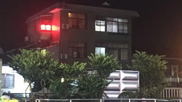 Un edificio de varios pisos con un letrero rojo a la izquierda, ventanas en los pisos superiores y una cerca en primer plano.