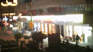 Vista nocturna de una calle con un escaparate muy iluminado y algunas personas caminando.
