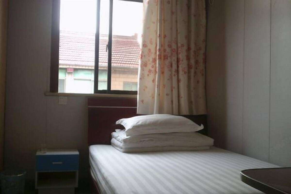 Ein Einzelbett mit weißer Bettwäsche, ein Fenster mit Vorhang, ein kleiner Nachttisch und ein Mülleimer.
