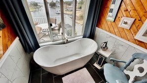 Een badkamer met een vrijstaand bad, een tafeltje met stoelen, een raam met uitzicht op een steiger en boten, en houten lambrisering aan de wanden.