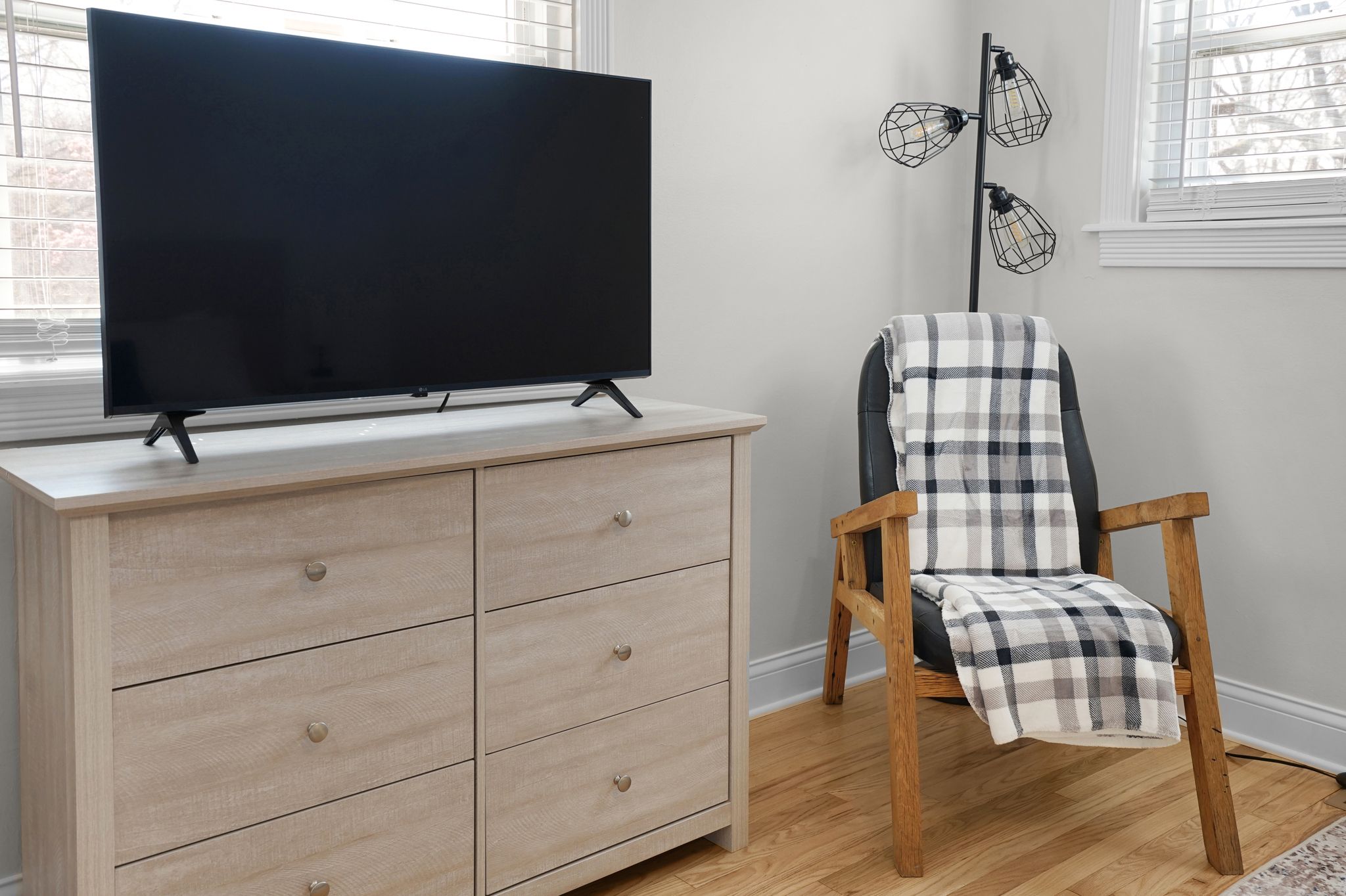 Una habitación con un televisor de pantalla plana grande sobre un aparador de madera clara, un sillón a cuadros con una manta y una lámpara de pie con tres luces.