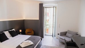 A modern hotel room with a large bed, a sofa, a desk, and a balcony with curtains.