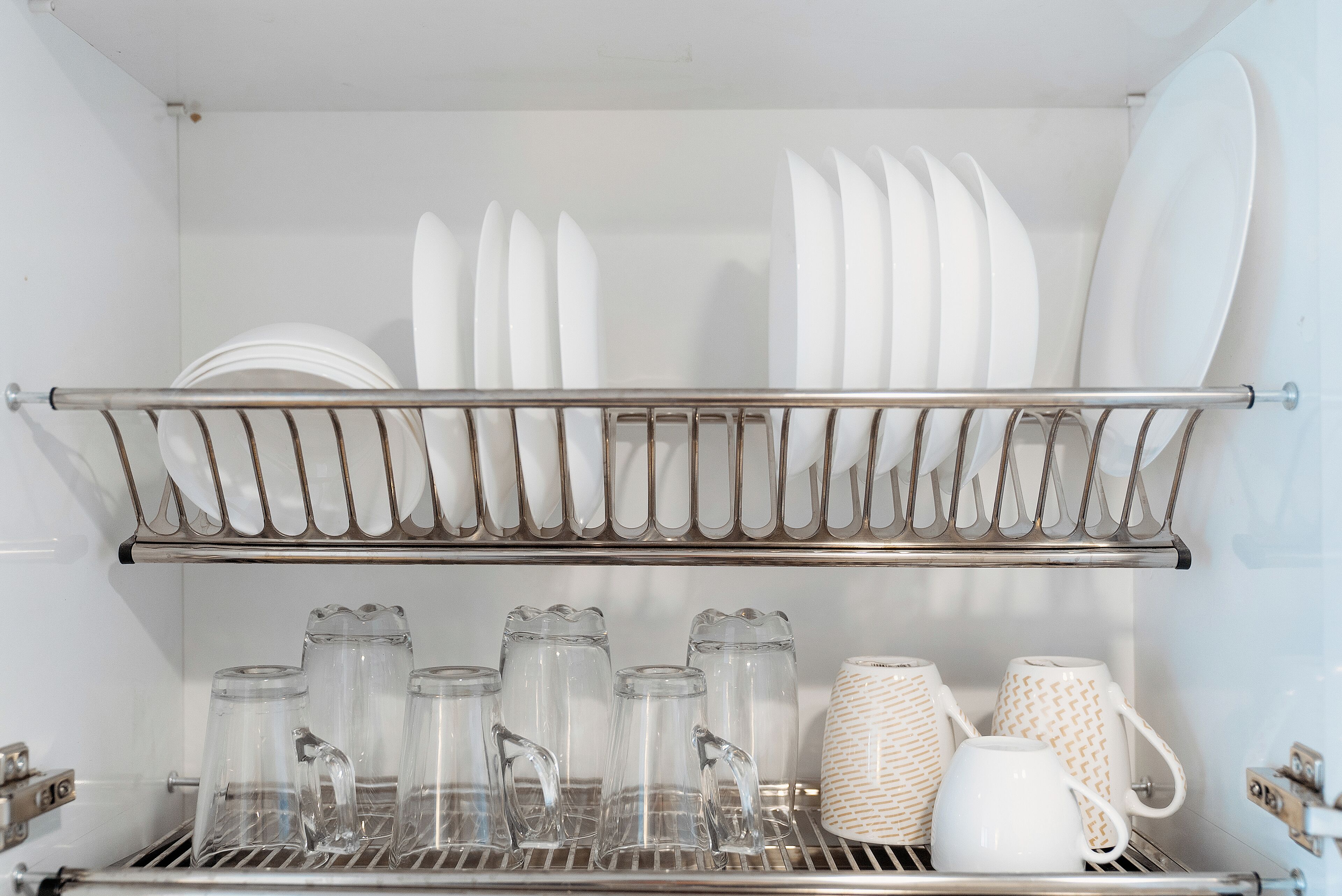Una cocina con un estante metálico que contiene platos y tazones blancos, y un repisa con jarras de vidrio transparente y una taza blanca.