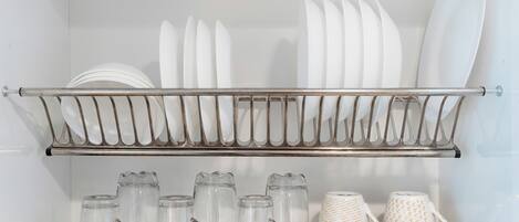 Una cocina con un estante metálico que contiene platos y tazones blancos, y un repisa con jarras de vidrio transparente y una taza blanca.