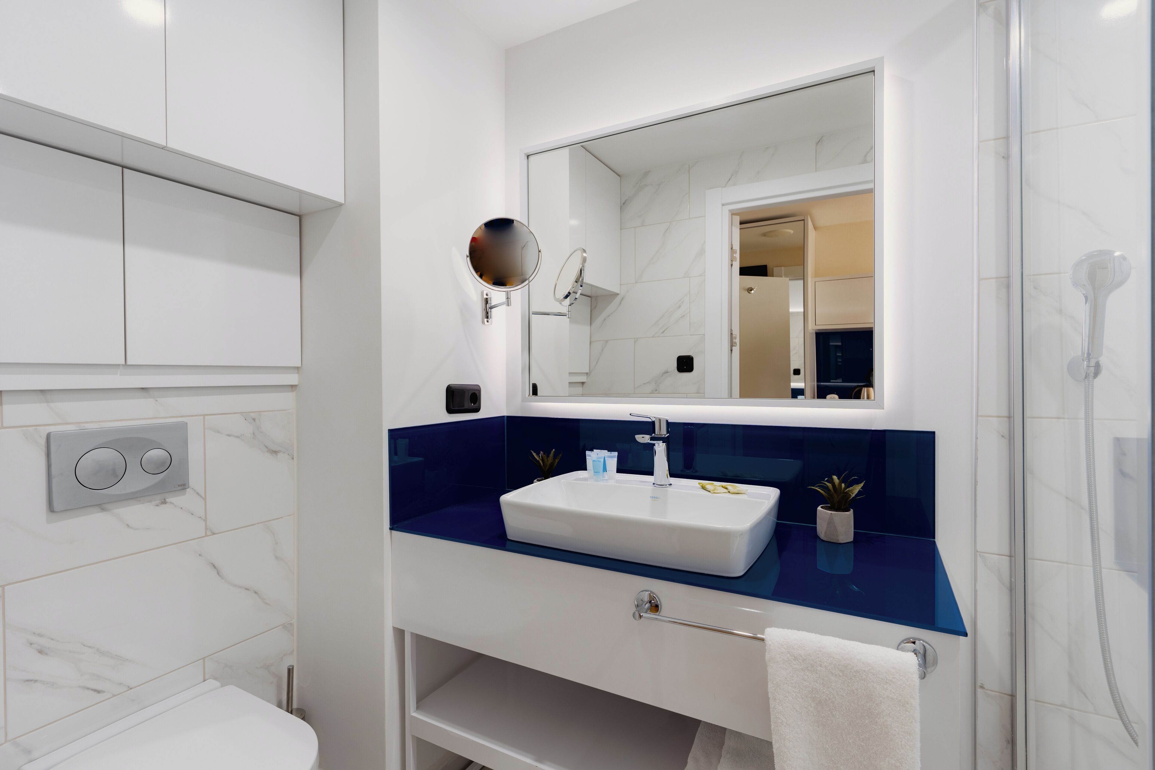 Ein modernes Badezimmer mit Toilette und Waschbecken in Weiß, einer blauen Arbeitsplatte, einem großen Spiegel und einer Duschkabine.