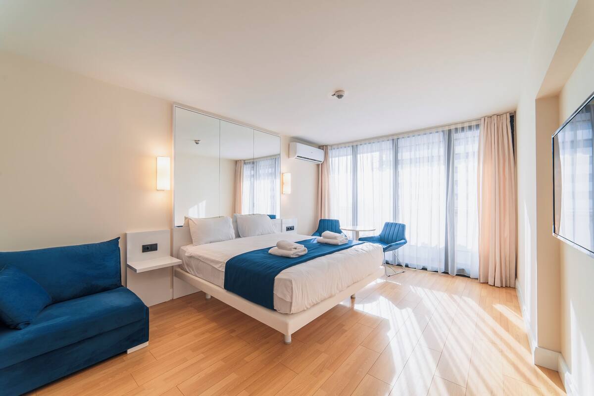 A modern hotel room with a large bed, a blue sofa, a desk, and a large window with curtains.