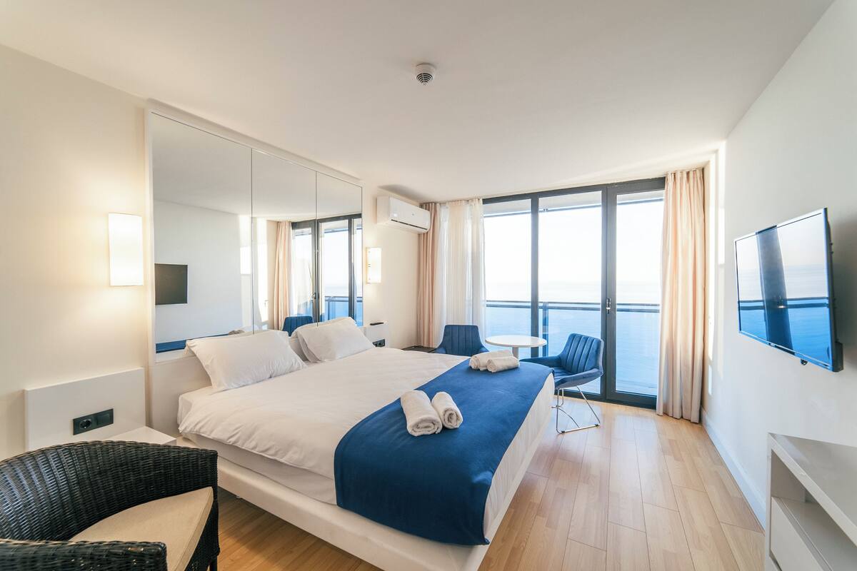 A modern hotel room with a large bed, a flat-screen TV, a balcony with a view, and a dining area.