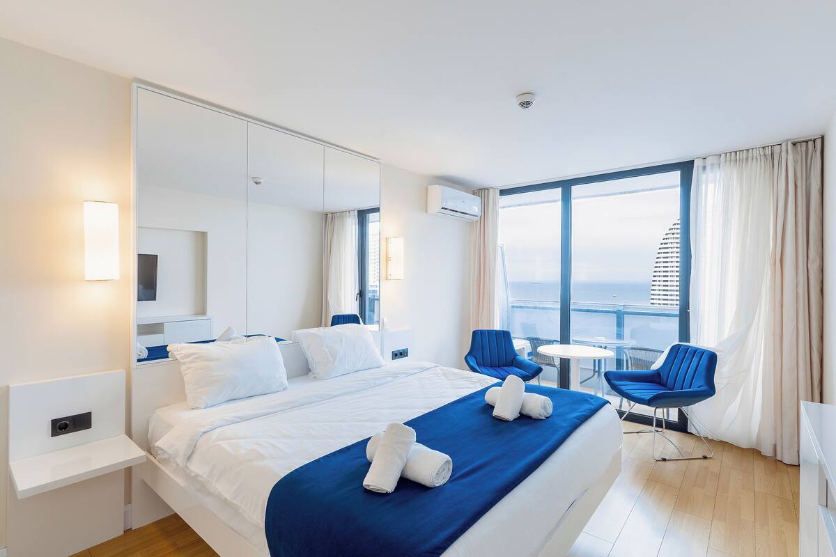 A modern hotel room with a large bed, a desk, a chair, and a view of the city.