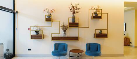 A modern lobby with two blue armchairs, a small round table, and geometric wall shelves with potted plants.