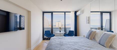Una habitació d'hotel moderna amb un llit gran, un televisor de pantalla plana al mur, un balcó amb vista a la ciutat i dues cadires blaves.