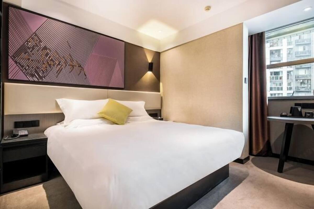A modern hotel room with a large bed, a desk, and a cityscape view.