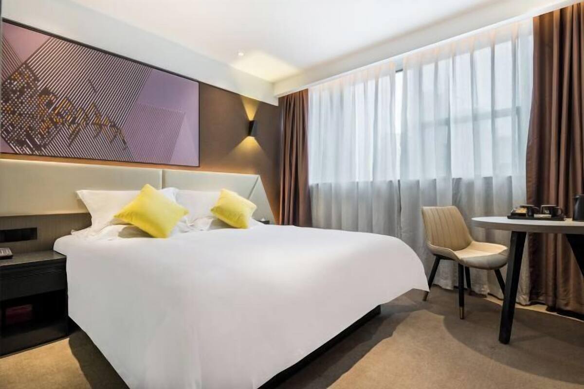 A hotel room with a large bed, a bedside table, a chair, a small table, and a large window with curtains.