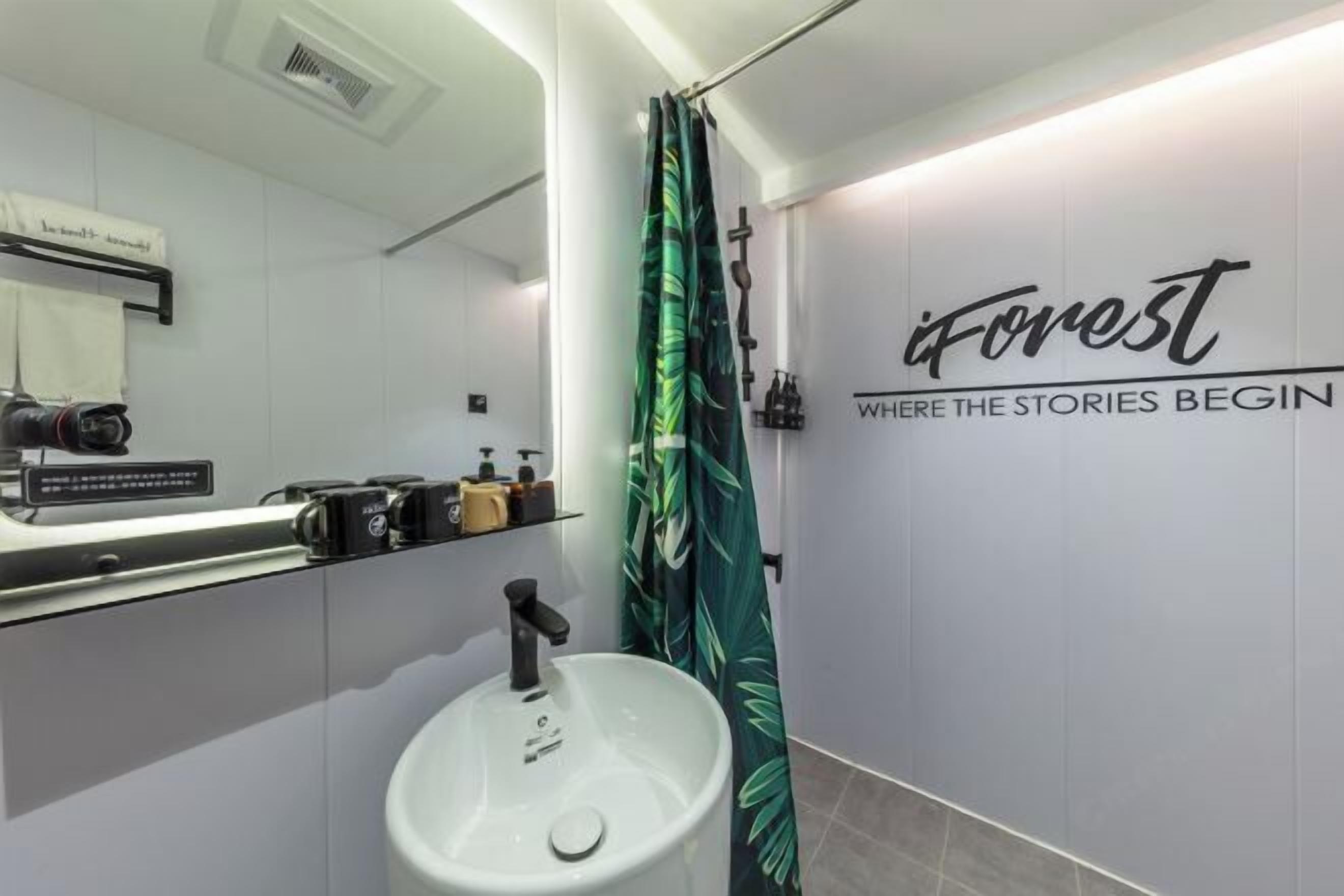 Un baño moderno con un lavamanos blanco, un espejo, una cortina de ducha con estampado de hojas y un cartel en la pared que dice 'iForest WHERE THE STORIES BEGIN'.